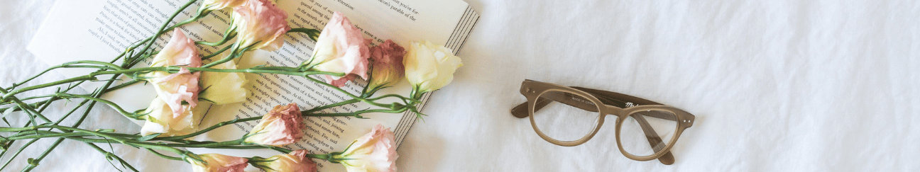 A long thin banner. A book and a pair of glasses lie on a wrinkled sheet. Yellow, white, and pink flowers have been laid over the book.