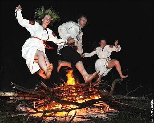 Three people dressed in ceremonial garb jump over the branches of a bonfire, a Litha tradition.