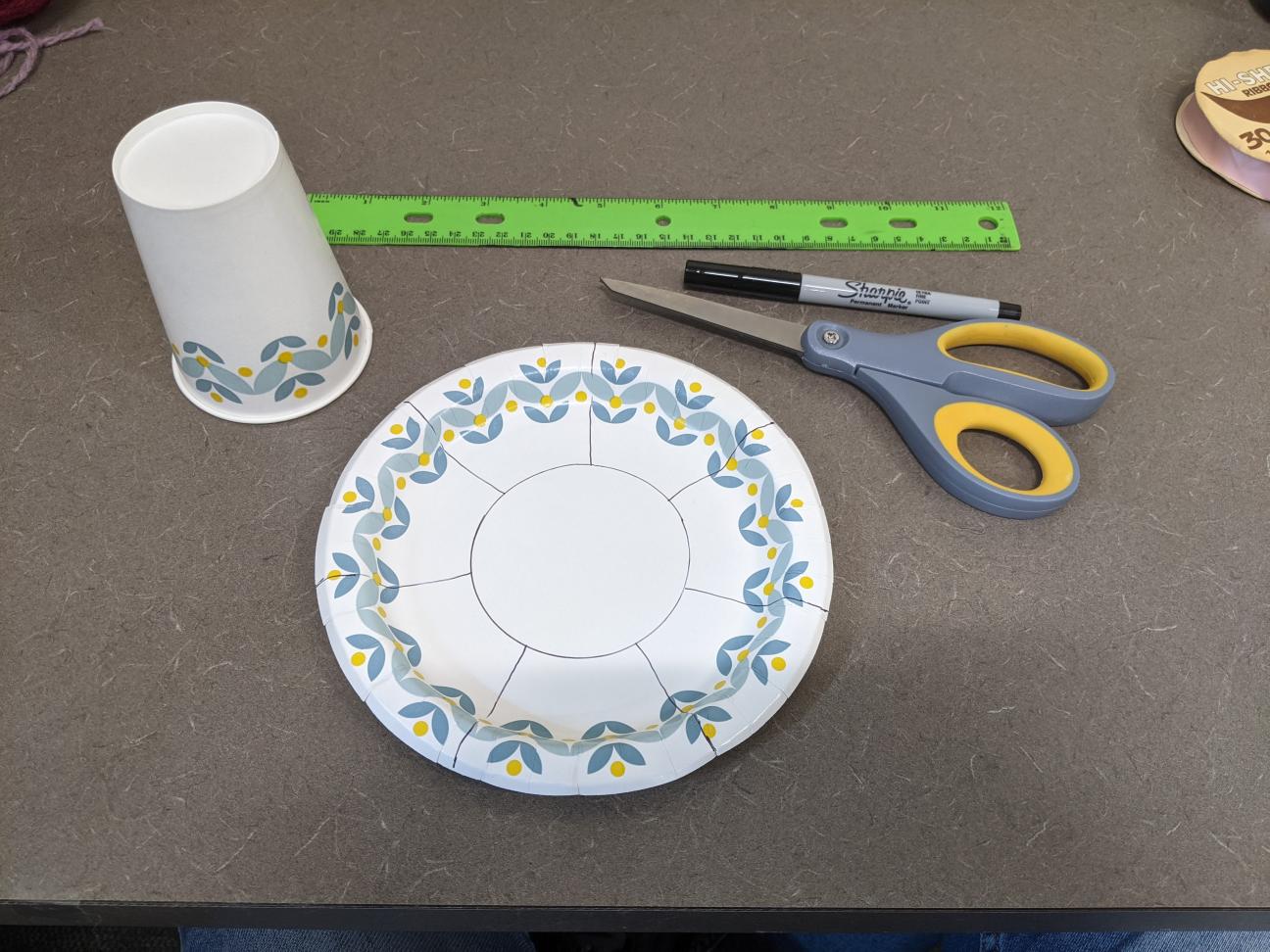 A paper plate and paper cup. The paper plate has a circle and lines drawn on it.