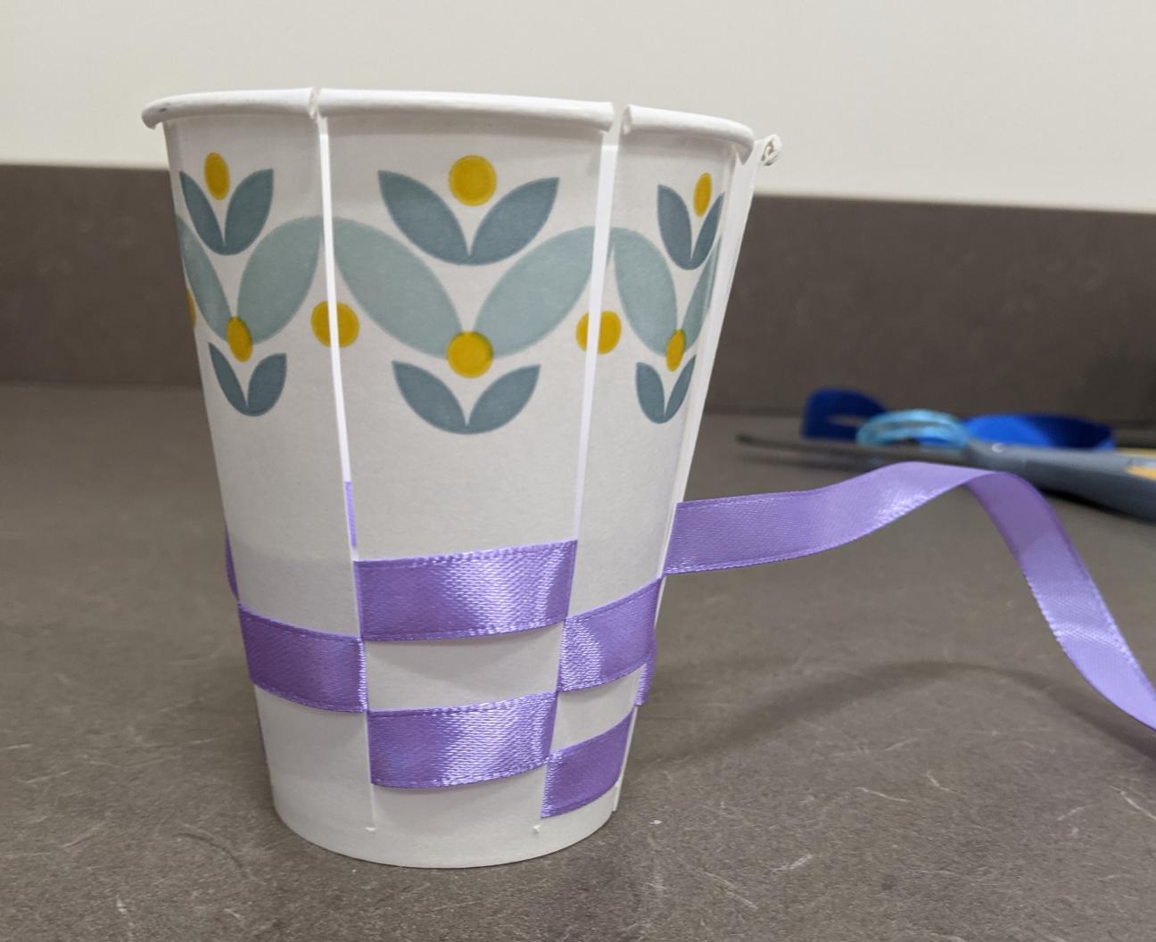 Weave in the ribbon. The ribbon pictured is purple and the cup is white with blue and yellow flowers at the top.