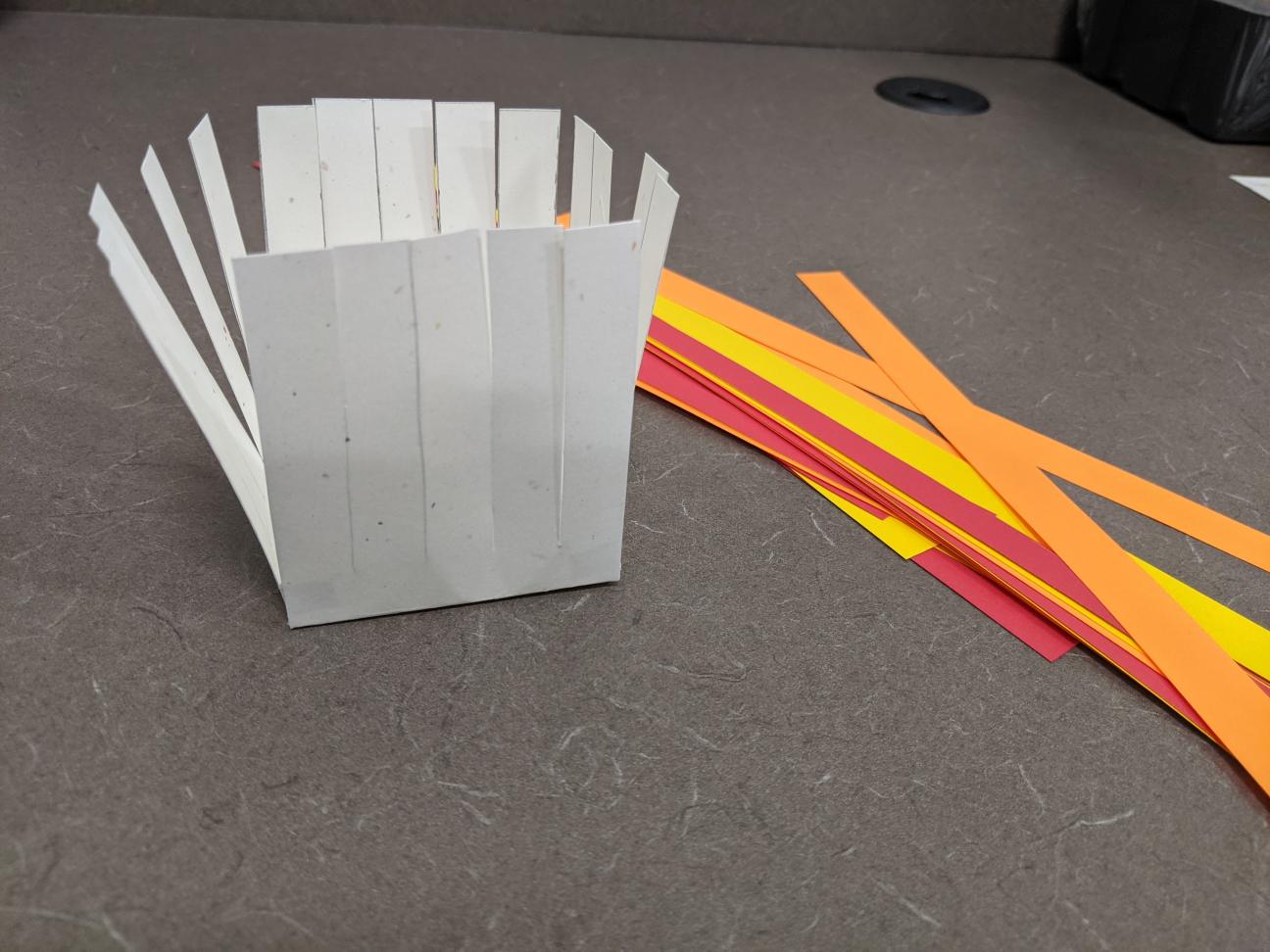 The glued together paper basket, now with thin strips of red, orange, and yellow paper next to it