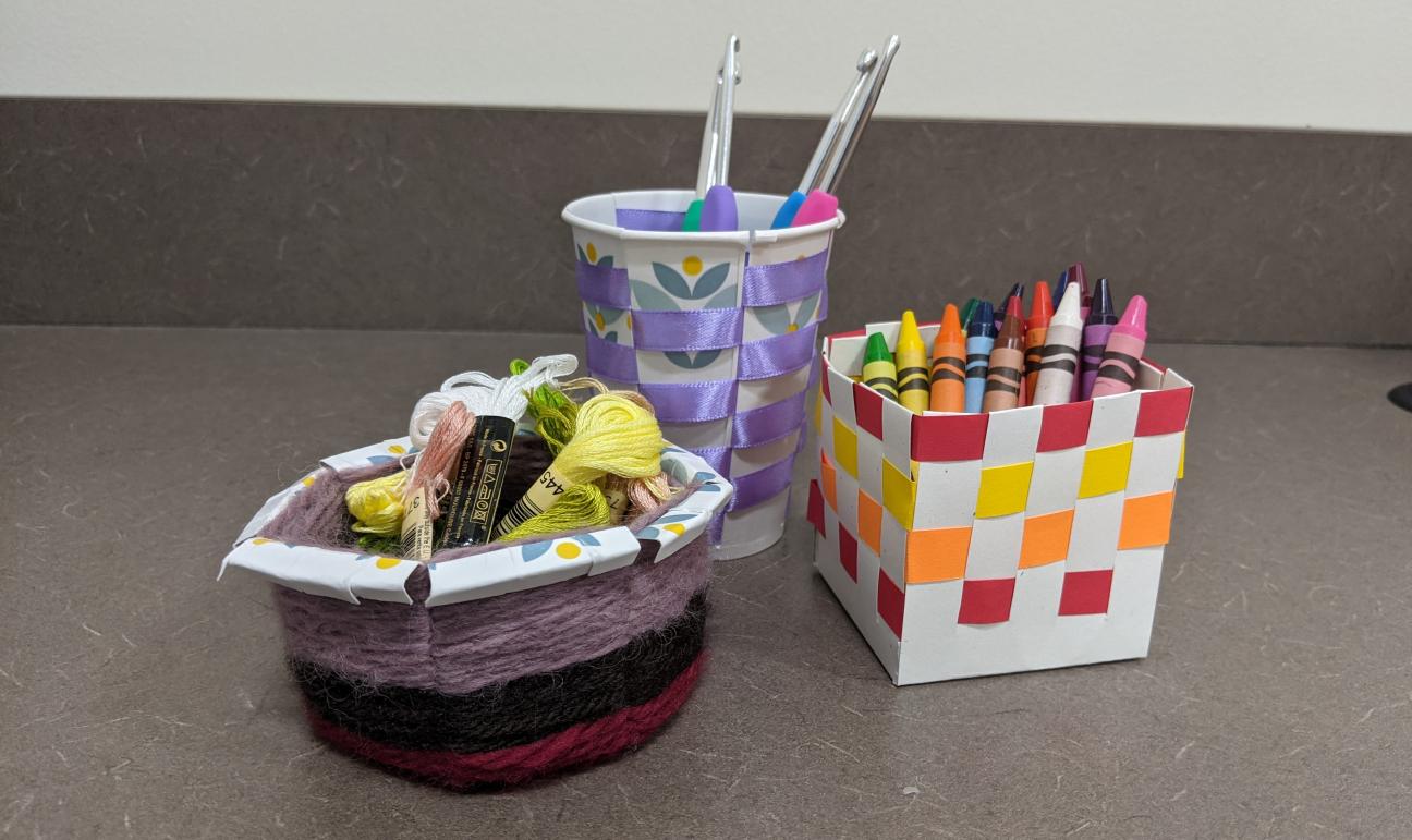 A picture of three baskets. One is made from a paper plate and yarn. Another is tall and made from a paper cup and ribbon. The last is a square and made with white and colored paper