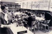 old black and white photo of inside the old library from 1956-1968 with a librarian holding up a card from the card catalog