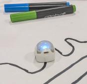 A small dome-shaped robot on a piece of paper next to two markers.