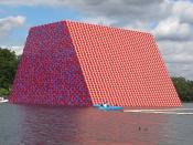 A barge of red, pink, blue, and white oil barrels bloats on the Serpentine